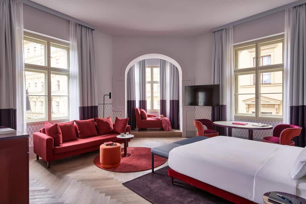 Quarto espaçoso do Falkensteiner Boutique Hotel Prague, com janelas grandes, sofá vermelho, poltrona, mesa redonda, cama e chão de madeira.
