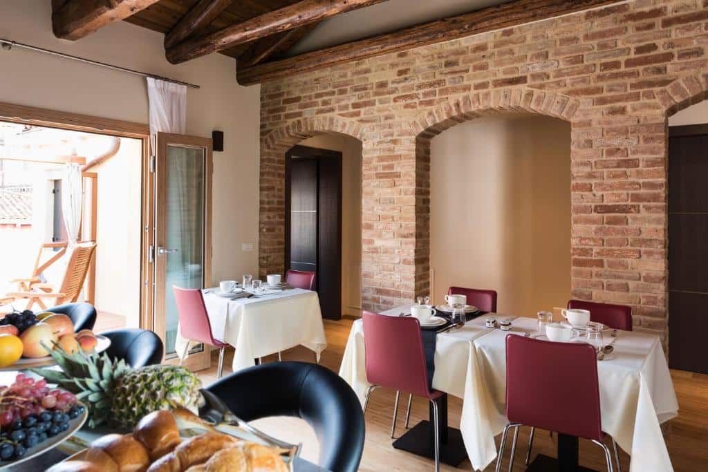 Sala de jantar rústica com paredes de tijolo à vista, mesas com toalhas brancas e cadeiras vermelhas, com refeição servida em pratos e taças, fruta e pão