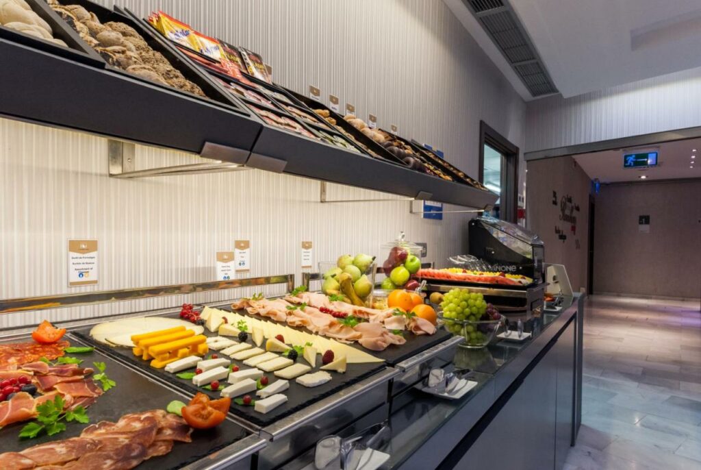 Buffet de pequeno-almoço do Ikonik Ramblas, com charcutaria, fruta e pães dispostos numa estação de dois níveis com pratos brancos.
