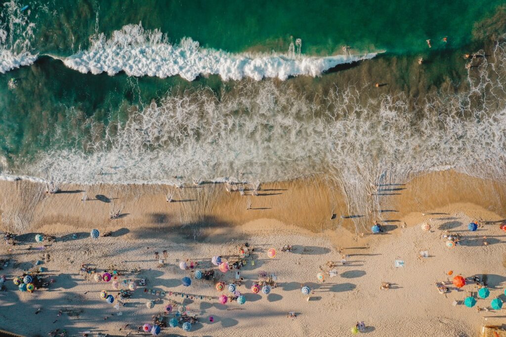 Vista aérea de uma praia movimentada com pessoas, chapéus de sol coloridos e ondas vindas do oceano.