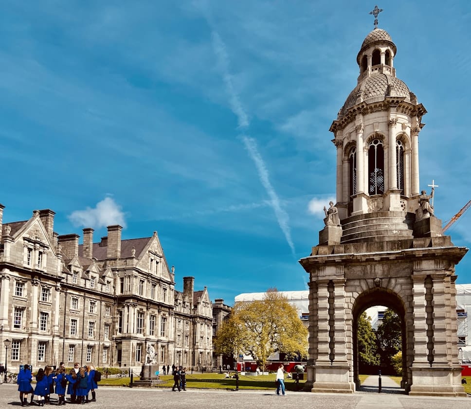 Imponente torre com campanário e cúpula ornamentada, em estilo arquitetónico europeu, erguida diante de edifícios históricos num campus universitário.