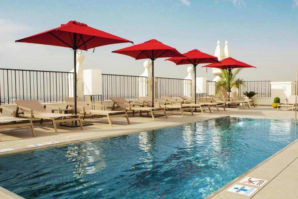 Piscina retangular no terraço com espreguiçadeiras e chapéus de sol quadrados e vermelhos do Riu Plaza España.