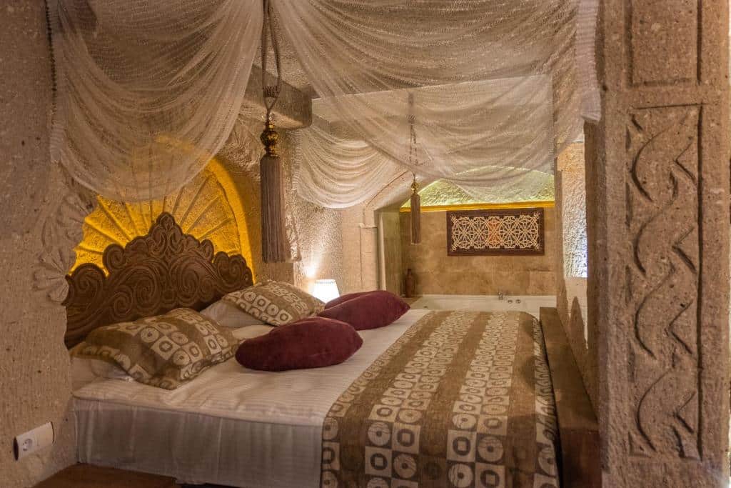 Cama do Grand Elite Cave Suites, um dos hotéis da Capadócia, com roupa de cama e almofadas estampadas sob um dossel transparente, num quarto de pedra com pormenores esculpidos.