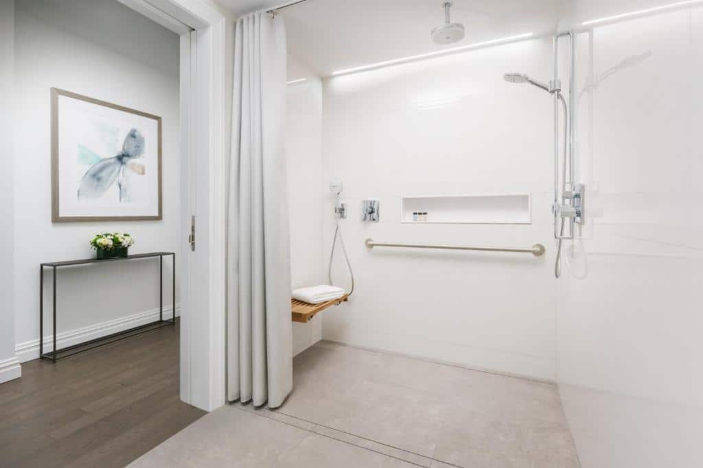 Casa de banho adaptada do The Wallace Hotel com um duche à direita e um banco de madeira rebatível na parede. Por cima, um nicho embutido com produtos de higiene; à esquerda, uma barra de apoio. À entrada, um cortinado branco.