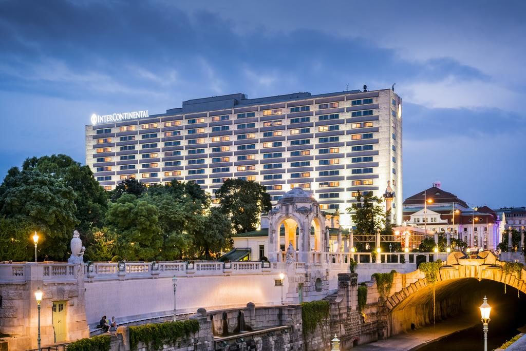 Edifício do InterContinental Wien ao anoitecer, com janelas iluminadas e a margem do rio com árvores e estruturas históricas de pedra.