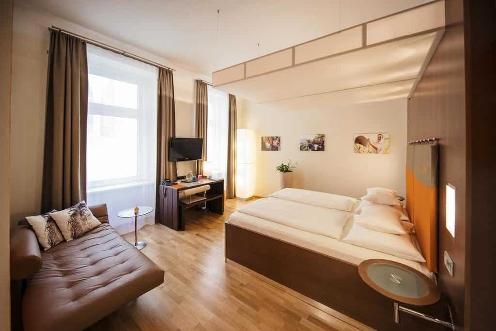 Quarto no Hotel Rathaus, com cama de casal, um sofá castanho, mesa com TV, chão de madeira clara e janelas grandes com cortinados.