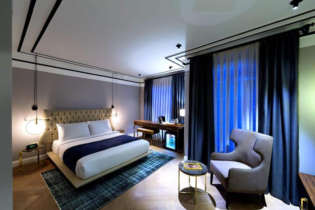 Quarto do Walton Hotels com uma cama de casal, secretária, poltrona, mesa redonda e cortinas escuras a cobrir grandes janelas.
