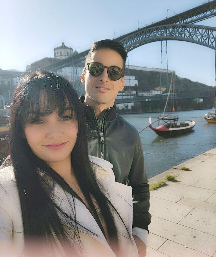 Casal a sorrir numa selfie com a Ponte Luís I ao fundo, no Porto, Portugal