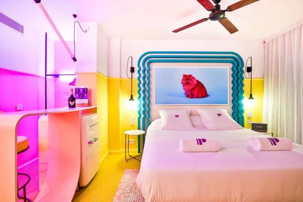 Quarto bem iluminado do Paradiso Ibiza Art Hotel com cama de casal, detalhes em néon, ventoinha de teto e um quadro de um gato vermelho por cima da cama.