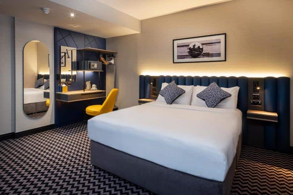 Quarto do Academy Plaza Hotel com cama de casal, cabeceira acolchoada azul, candeeiro de parede e espelho circular. Estrutura decorativa de madeira preta atrás da cabeceira e pavimento com padrão geométrico a preto e branco.