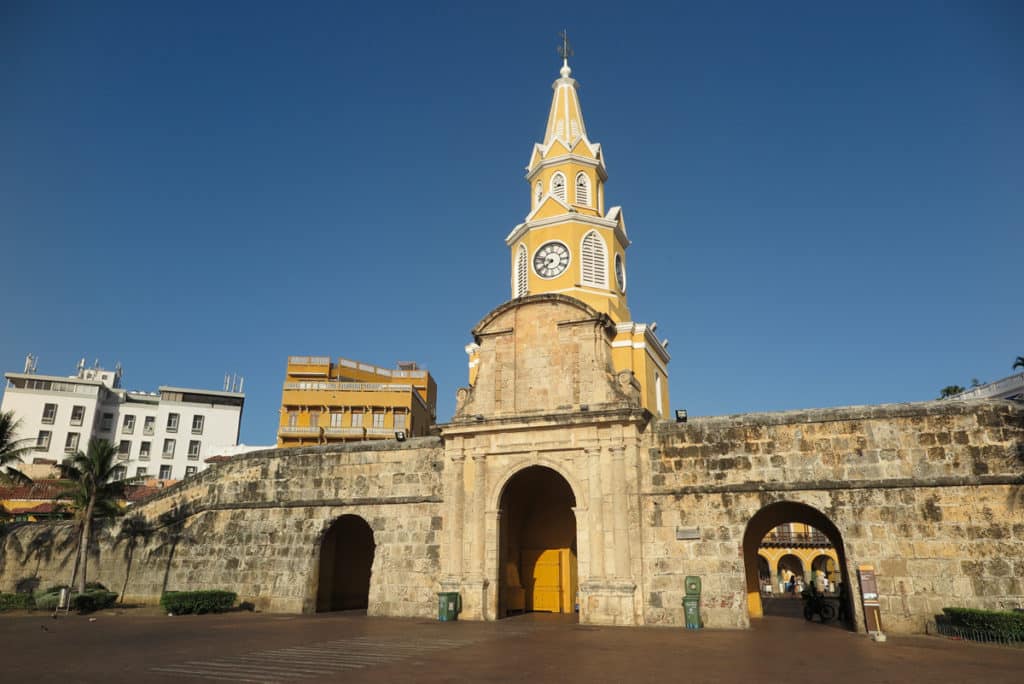 Entrada monumental da Cidade Amuralhada de Cartagena, com um grande arco de pedra encimado por uma torre amarela com relógio, contra o céu azul.