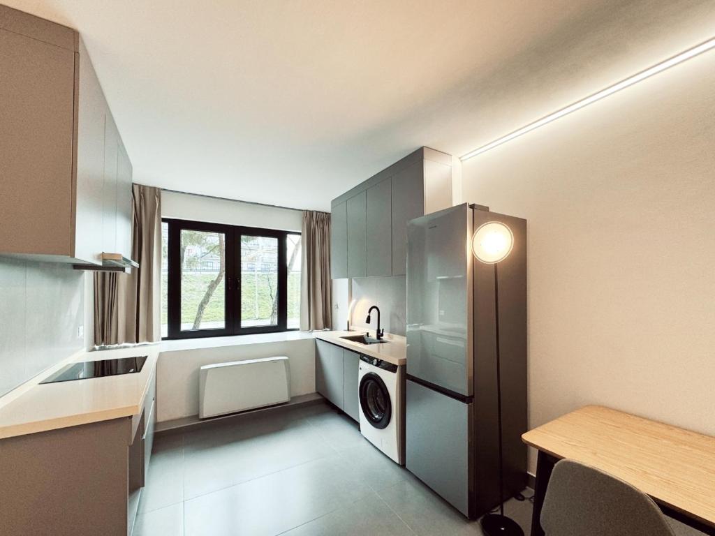Cozinha moderna com eletrodomésticos e utensílios em cinzento e branco, incluindo frigorífico e máquina de lavar roupa, bancada de madeira clara e janela com vista para o exterior