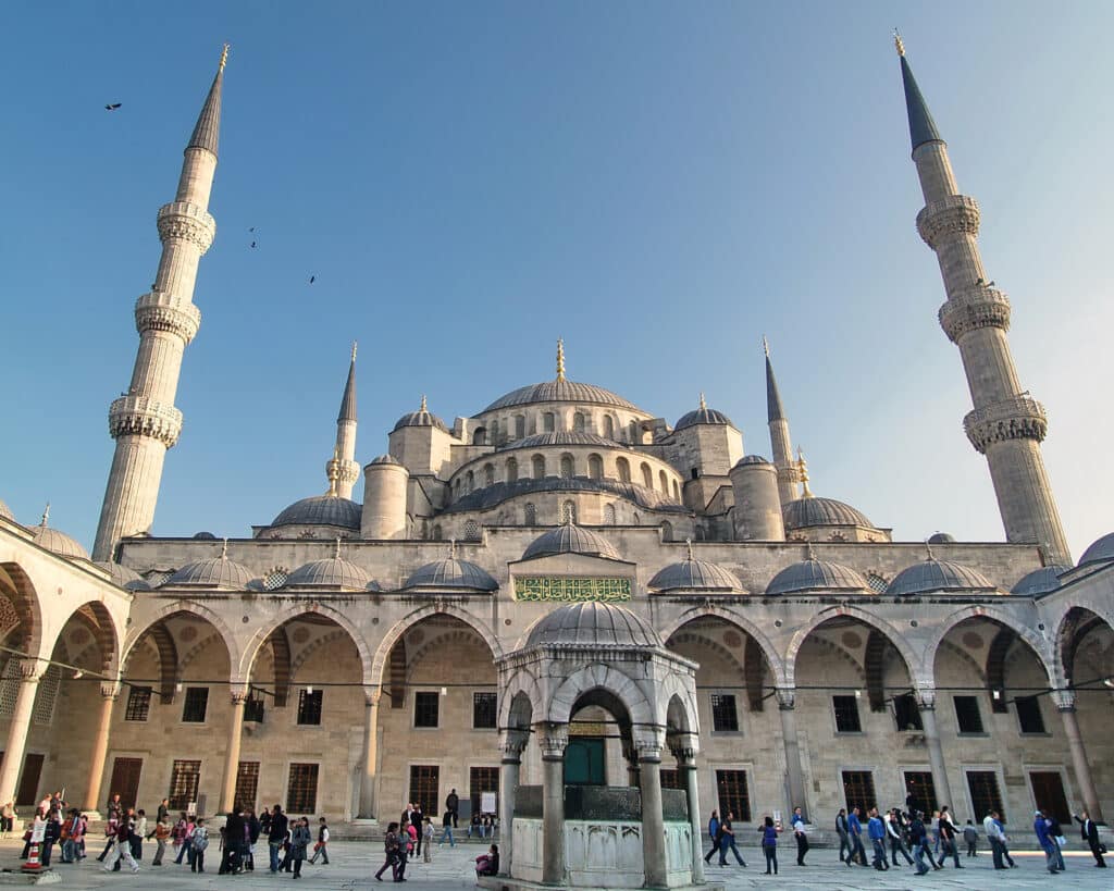 Pessoas caminham em frente à Mesquita Azul em Istambul, com cúpulas, arcos e quatro minaretes altos num céu claro.