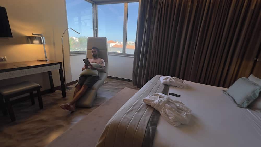 Uma pessoa sentada numa espreguiçadeira num quarto com cama grande, secretária e janela com vista para a cidade, no Neya Lisboa Hotel.