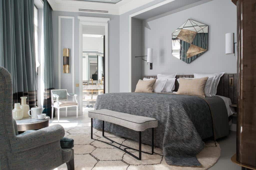 Quarto do Nolinski Paris com uma cama grande, roupa de cama cinzenta, espelho geométrico, banco, poltrona e detalhes decorativos.