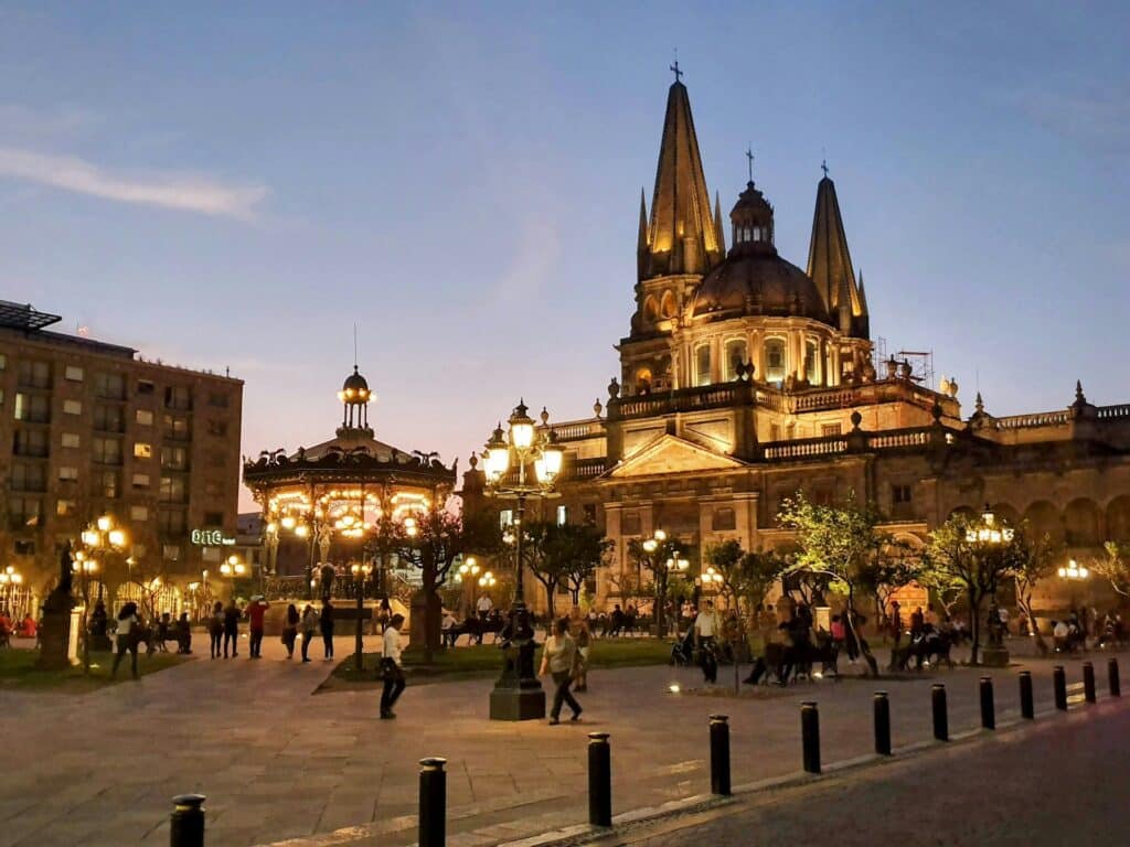 Praça da Catedral Metropolitana de Guadalajara à noite, com cúpula e torres iluminadas contra o céu escuro, rodeada de pessoas a passear.
