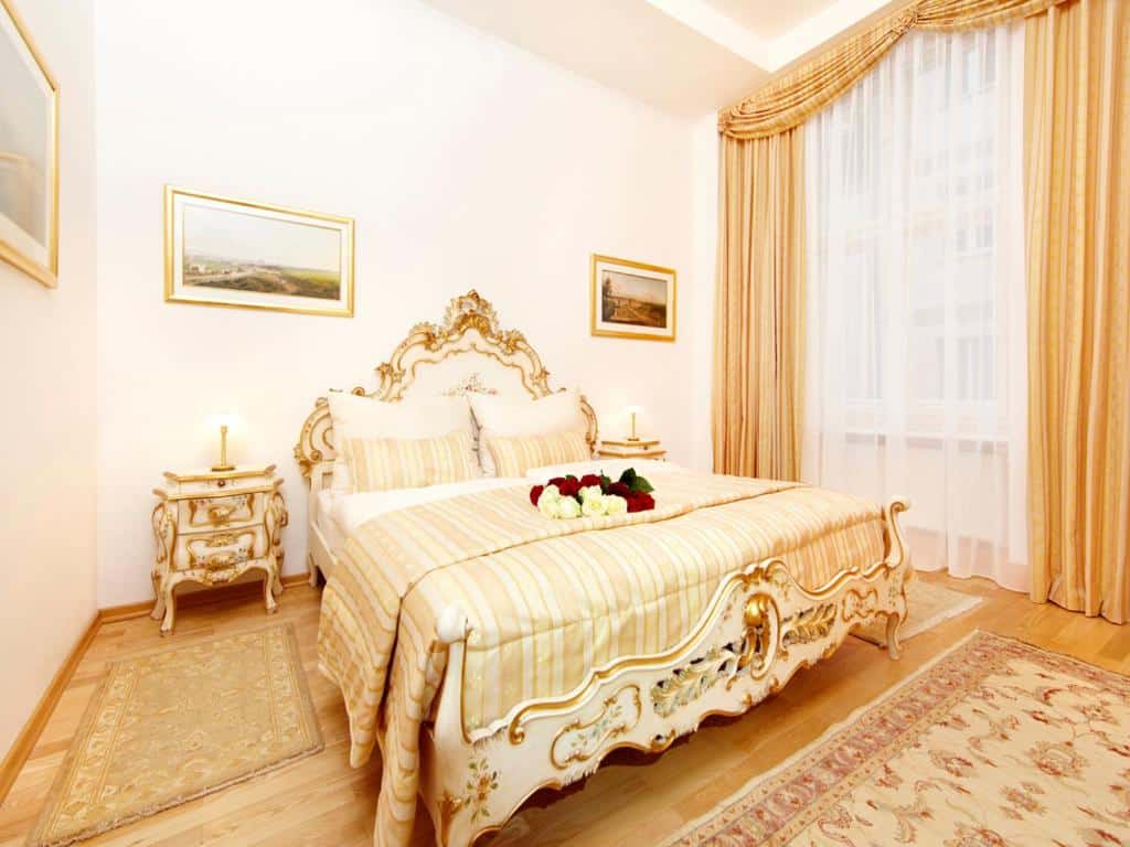 Quarto ornamentado no Kaiserin Sisi, com uma cama esculpida, mobiliário com apontamentos dourados, roupa de cama às riscas e uma grande janela com cortinados dourados.