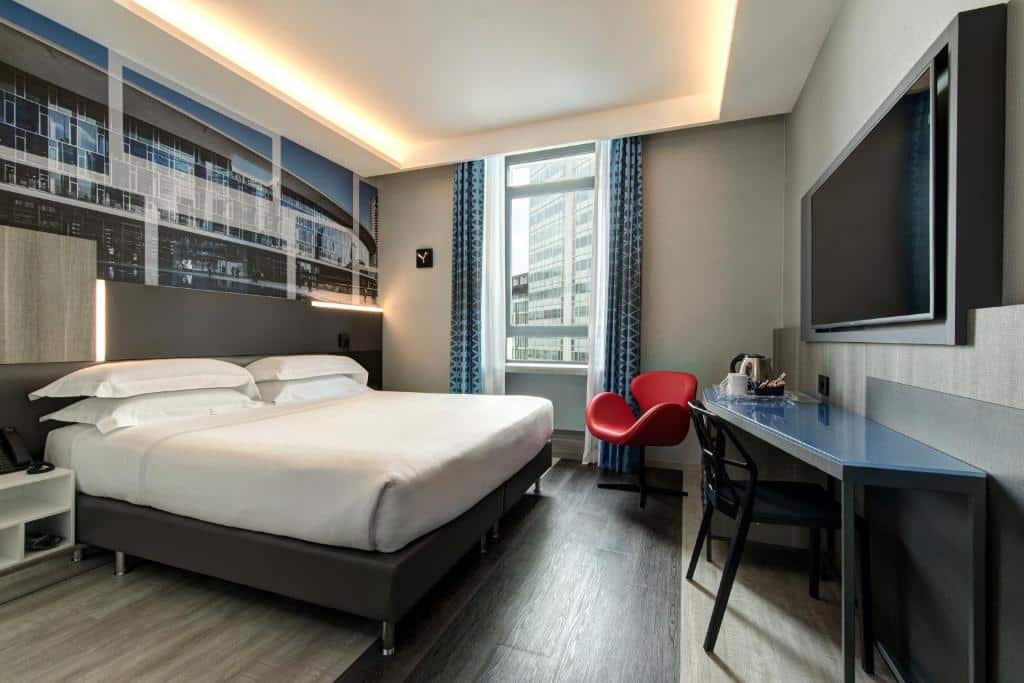 Quarto no iQ Hotel Milano com janela com cortinas, cama de casal, secretária com cadeira, televisão e uma poltrona vermelha