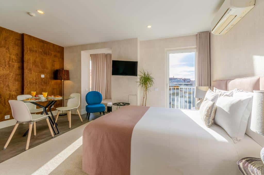 Quarto do LX51 Studios & Suites com uma pequena varanda com cortinas, uma cama de casal, uma televisão, uma mesa de vidro com três cadeiras, uma poltrona e um candeeiro de pé.