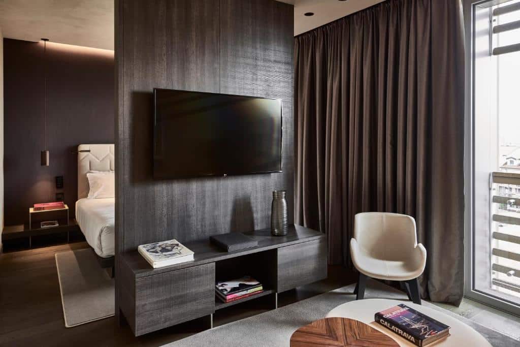 Quarto dividido entre a cama e um painel com televisão e, do outro lado, mesa e cadeiras no Hotel VIU Milan