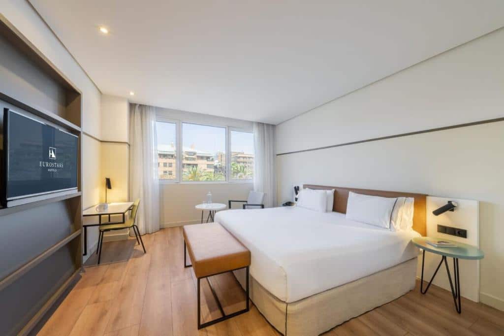 Quarto do Eurostars Acteón com cama de casal, TV de ecrã plano, móveis de madeira e janelas com vista para os edifícios da cidade.