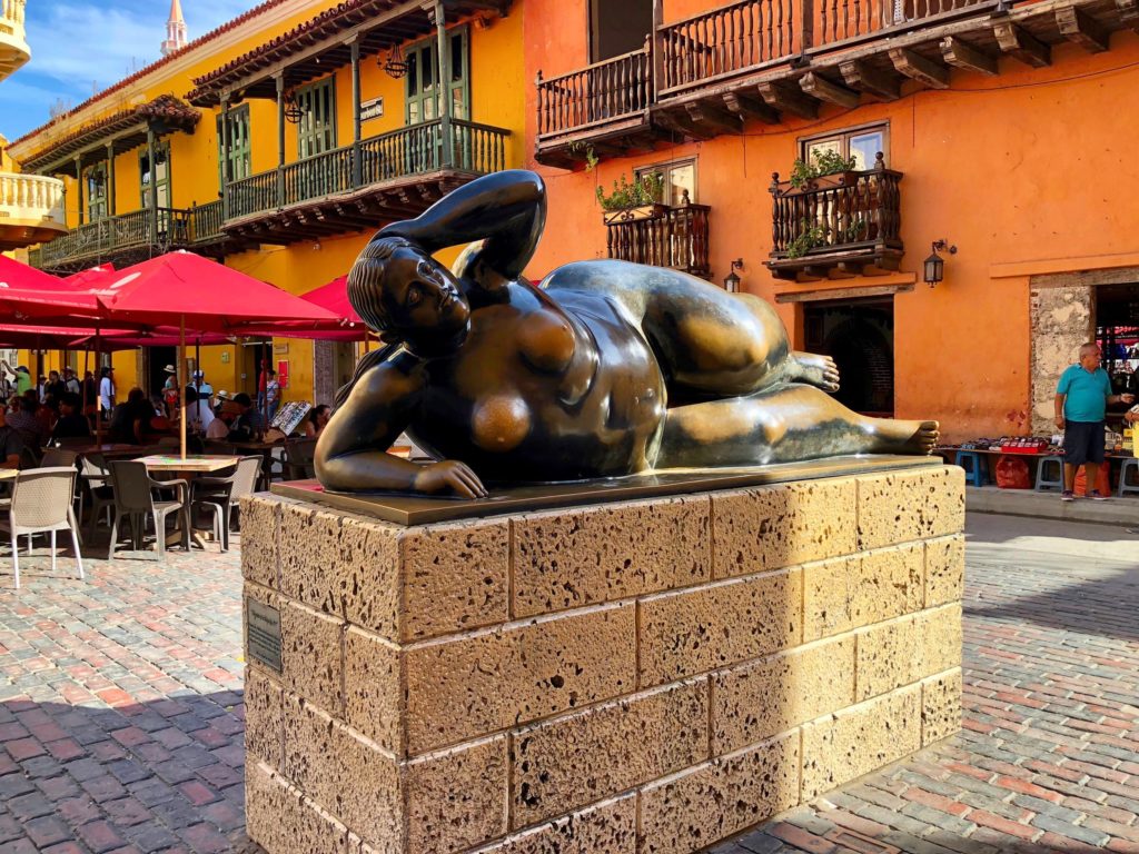 Escultura em bronze de uma mulher deitada, de Fernando Botero, na Praça de Santo Domingo, em Cartagena, rodeada de edifícios coloniais coloridos.
