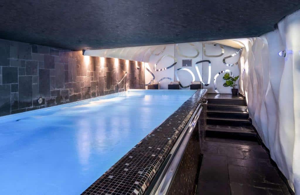 Piscina interior do Anantara New York Palace com água azul, pavimento de azulejos pretos, paredes de pedra e decoração branca moderna com recortes circulares.