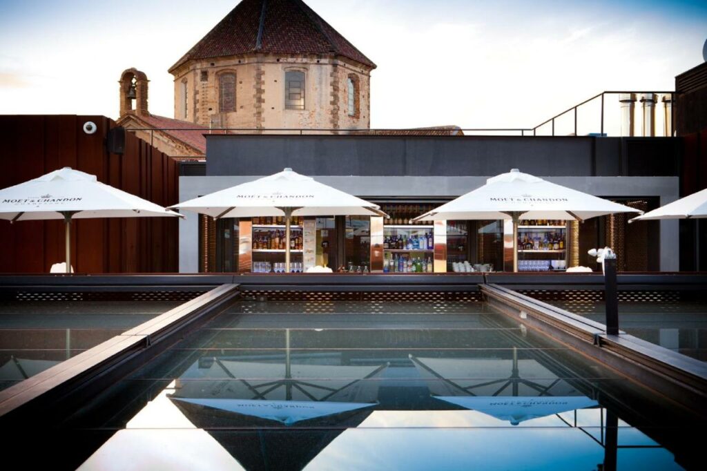 Piscina do Hotel España Ramblas, com chapéus de sol brancos ao longo da extensão da água e o edifício do hotel ao fundo.