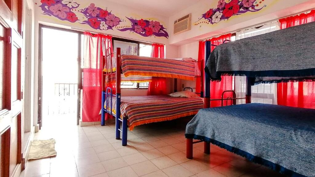 Um quarto do Hostel Vallarta bem iluminado com dois beliches, cortinas vermelhas, pavimento de ladrilho e mural floral por cima das janelas e da porta.
