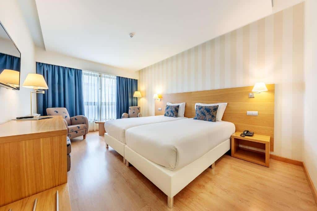 Quarto no Steyler Fátima Hotel Congress & Spa com duas camas de solteiro, mobiliário de madeira, duas poltronas, cortinas azuis e iluminação suave, para representar onde ficar em Fátima.