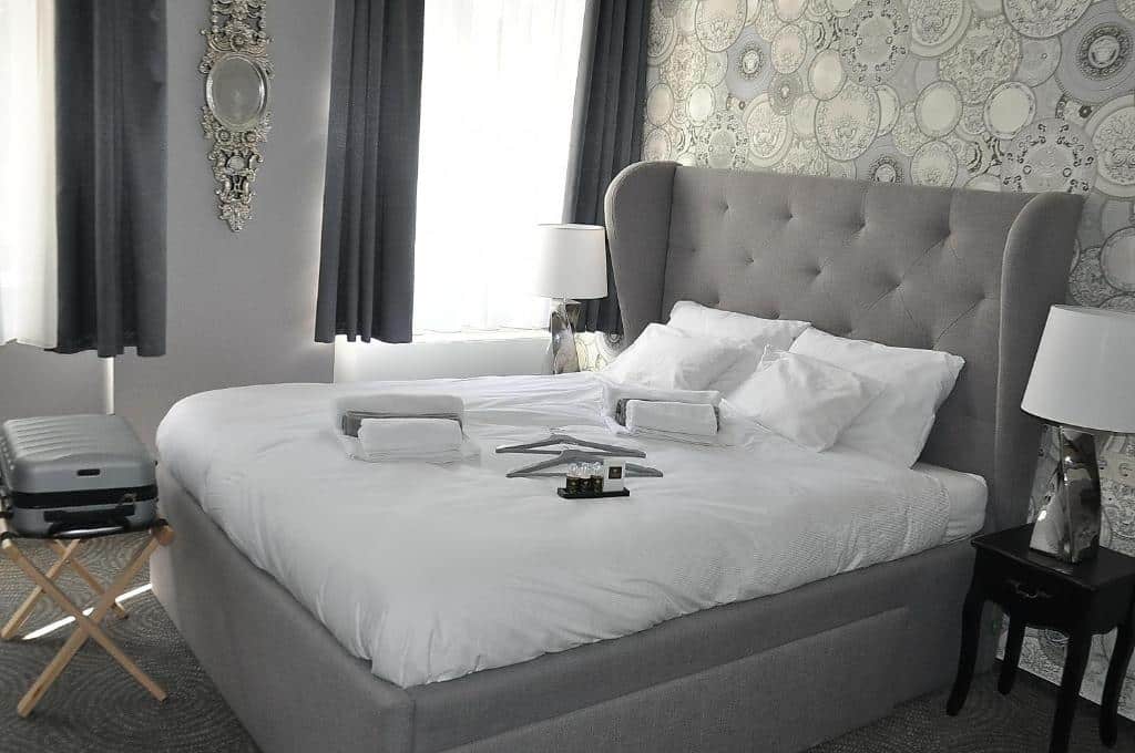 Uma cama bem arrumada do Váralja Home, com roupa de cama branca, num quarto de tema cinzento, com mala num suporte e candeeiros nas mesas de cabeceira.