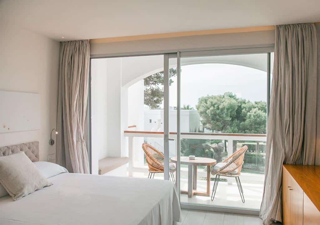 Quarto bem iluminado do Destino Pacha Ibiza com cama, cortinas claras e varanda com duas cadeiras e uma pequena mesa com vista para a vegetação.