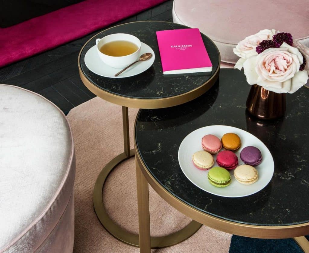 Duas mesas redondas no Fauchon L’Hôtel Paris exibem uma chávena de chá, um livro cor-de-rosa, um vaso de flores e um prato de macarons coloridos. Este é um dos hotéis de luxo em Paris.
