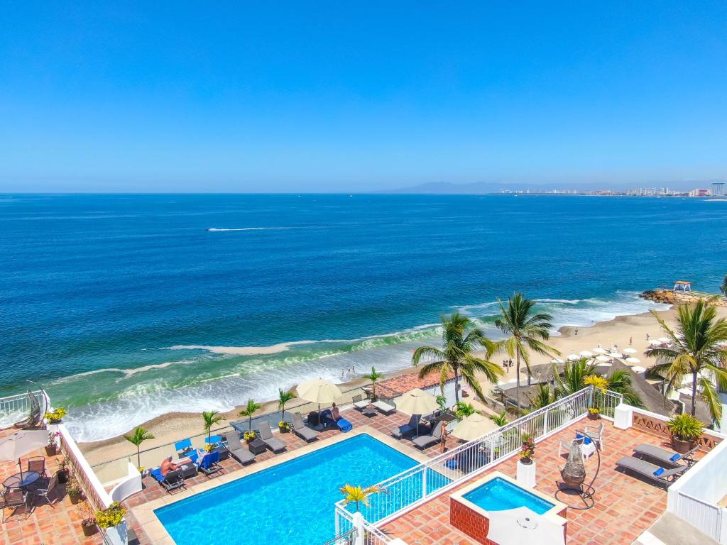Um resort à beira-mar com piscina, espreguiçadeiras, palmeiras e uma praia de areia com vista para um oceano azul calmo sob um céu claro.