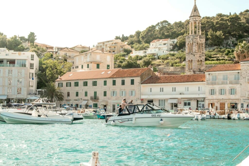 Barcos atracados num porto ensolarado de Hvar Town, com edifícios e uma igreja de torre alta ao fundo.
