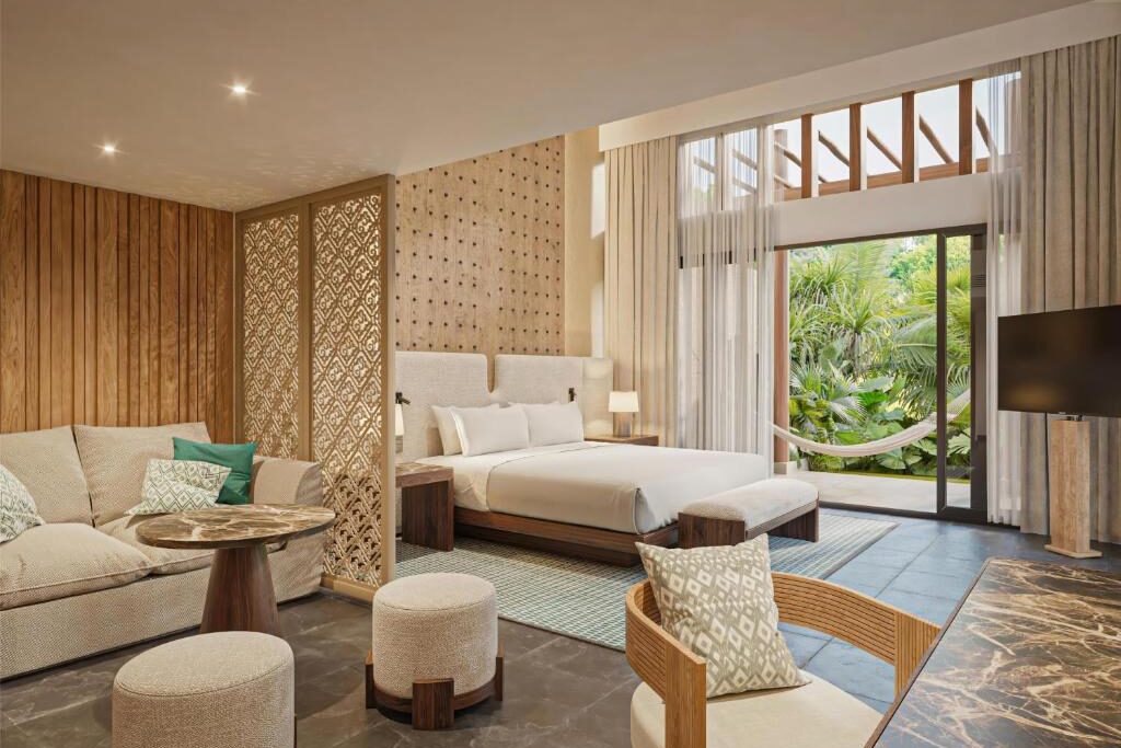 Quarto do Banyan Tree Mayakoba com cama king size, decoração em madeira e vista para área exterior com rede. Em primeiro plano, uma zona separada do quarto com sofá e poltronas.