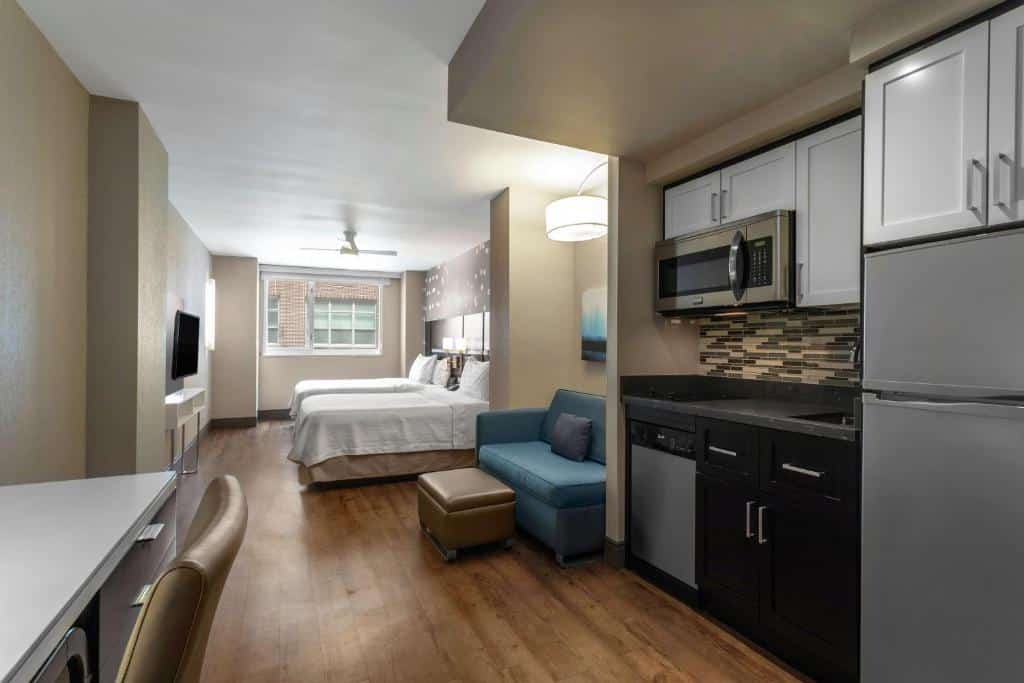 Na suíte do Homewood Suites Midtown Manhattan Times Square South há duas camas de casal com cabeceiras estofadas ao centro. À esquerda, uma pequena cozinha com armários brancos, micro-ondas e lava-loiça; à direita, uma zona de estar com sofá azul e mesa de centro. Ao fundo, uma janela grande com cortinados.