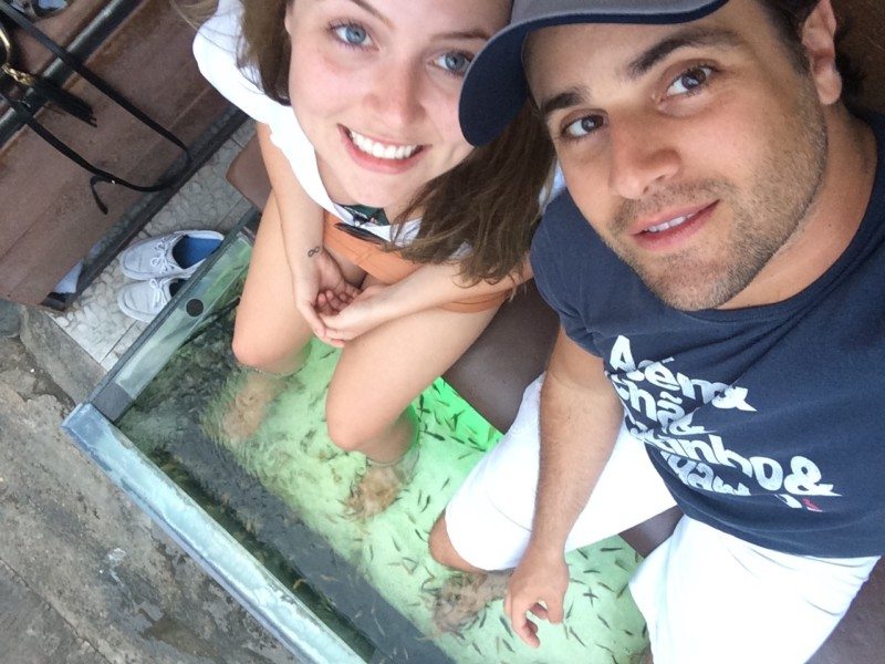 Pessoas com os pés submersos num tanque de vidro cheio de pequenos peixes, numa sessão de pedicure com peixes.