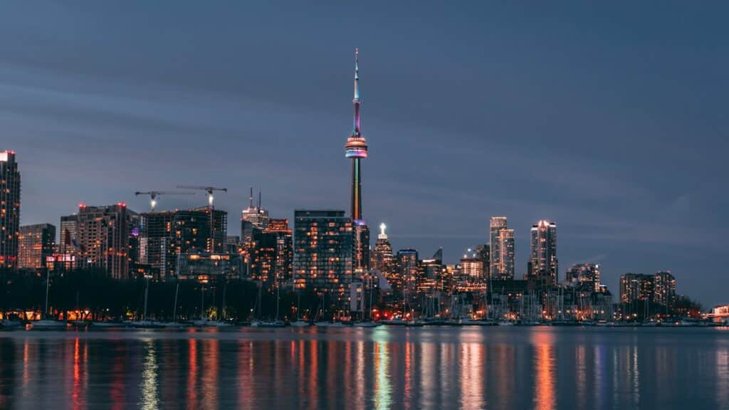 Skyline iluminado de Toronto à noite, com a emblemática CN Tower a destacar-se ao centro e barcos atracados a refletir a paisagem urbana na água calma do lago