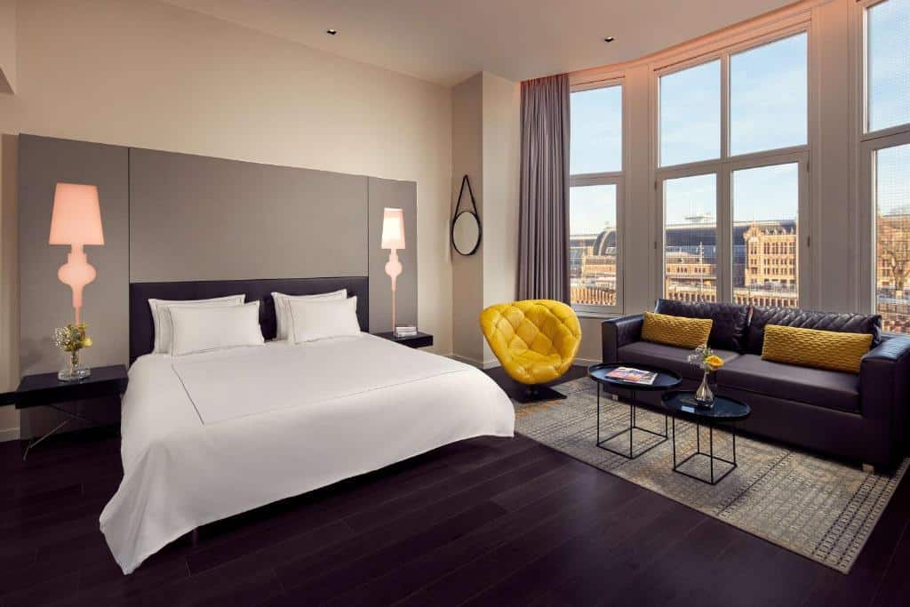 Quarto moderno do Park Plaza Victoria Amsterdam, que ilustra onde ficar em Amesterdão: cama grande, poltrona amarela, sofá cinzento, mesas de apoio e janelas grandes com vista para a cidade.