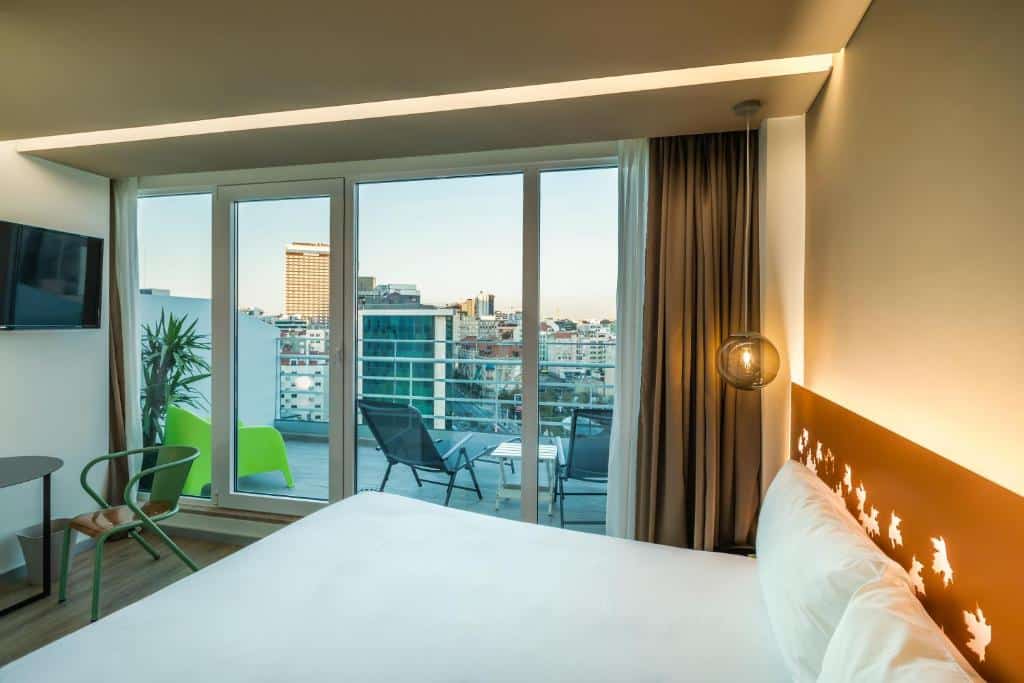 Quarto do Hotel Ibis Styles Lisboa Marquês de Pombal com varanda com cadeiras e vista da cidade; no quarto há uma cama de casal, uma televisão e uma mesinha com uma cadeira