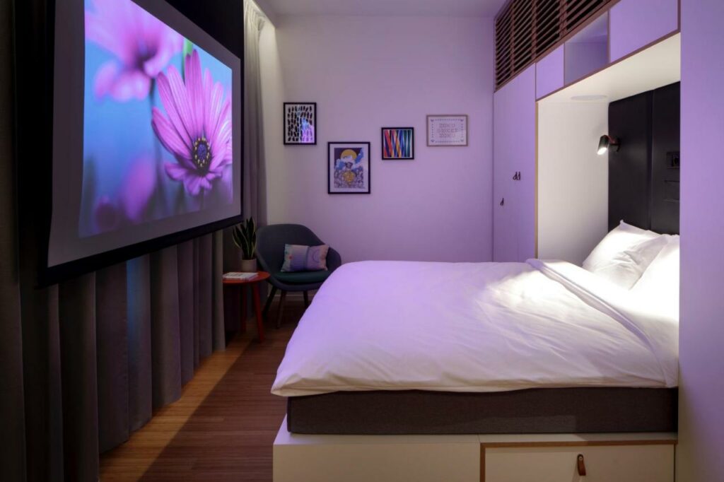 Quarto do hotel Zoku Copenhagen. Tem uma cama de casal, armário integrado na parede, uma cadeira e um ecrã de projeção à frente da cortina, com iluminação roxa ambiente.
