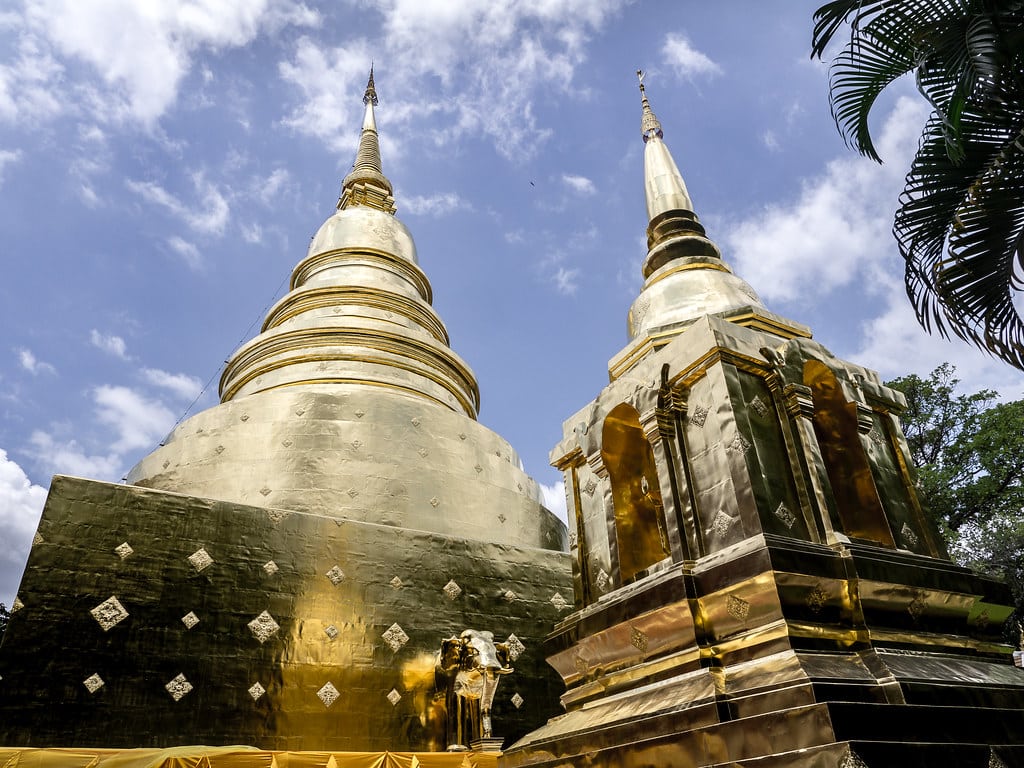 Pagodes dourados com desenhos intrincados rodeados de vegetação, sob céu azul, no Wat Phra Singh, em Chiang Mai.