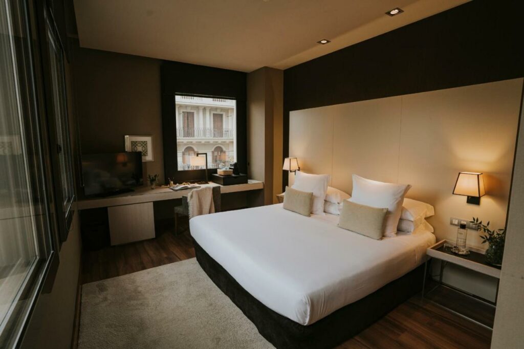 Quarto do Grand Hotel Central. Uma cama de casal com candeeiros nas mesas de cabeceira, tapete aos pés e uma bancada com televisão ao fundo, junto a várias janelas.