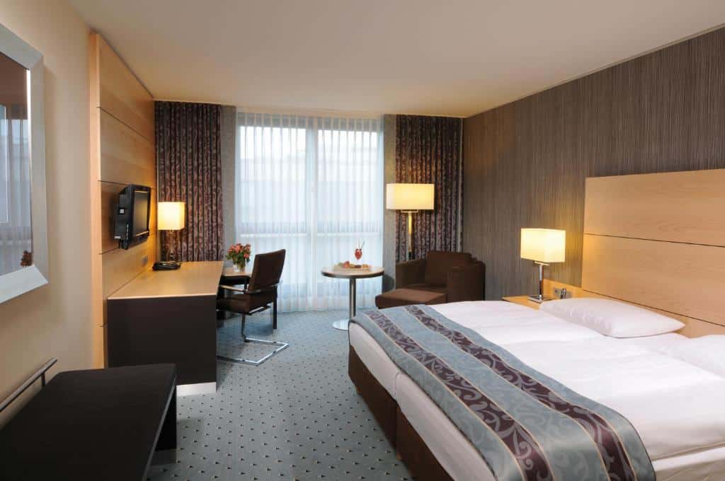 Quarto do Maritim Hotel Düsseldorf com cama de casal, poltrona com mesinha redonda e secretária com TV. Representa hotéis em Dusseldorf.
