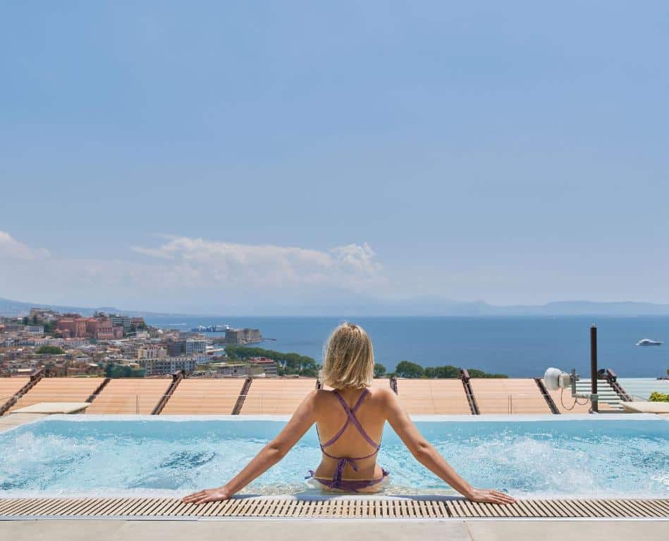 Mulher sentada na borda de uma piscina a observar a vista de uma cidade litoral com mar azul e montanhas ao fundo, no De Bonart Naples.