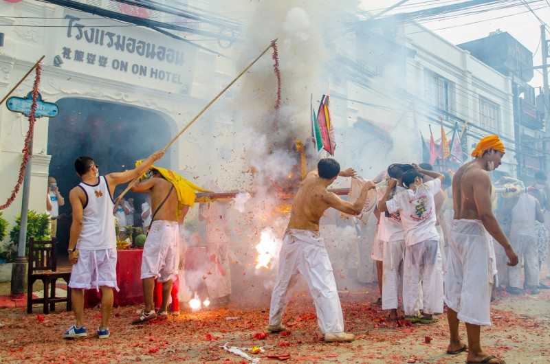 Participantes vestidos de branco com bandeiras durante um festival em Phuket, com fumo e fogo de artifício.