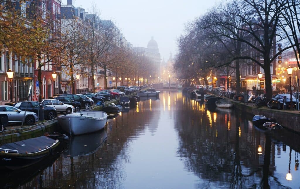 Canal de Amesterdão, um dos destinos mais visitados da Europa, com barquinhos parados nas margens e ruas em ambos os lados, onde há carros estacionados, árvores e edifícios coloridos