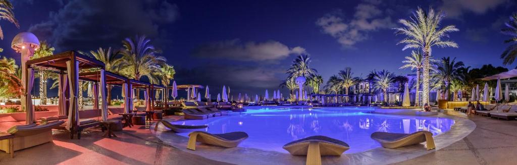 Vista noturna da zona da piscina do Destino Pacha Ibiza, com espreguiçadeiras, cabanas, palmeiras e iluminação suave.