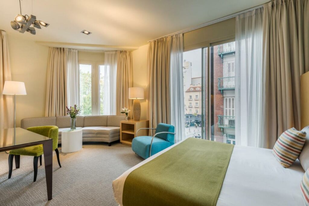 Suíte do Room Mate Alicia, um dos melhores hotéis em Madrid. Uma cama de casal fica em frente à zona de estar, composta por um sofá e uma poltrona com mesinhas com flores e candeeiros. Na parede esquerda há uma secretária com cadeira estofada. Uma ampla janela de vidro na parede direita tem vista para a cidade.